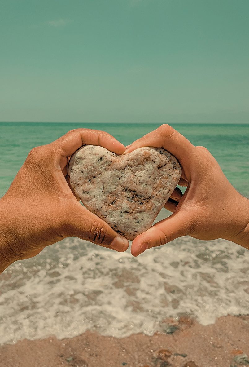 Hands shaping a heart with a stone, ocean behind